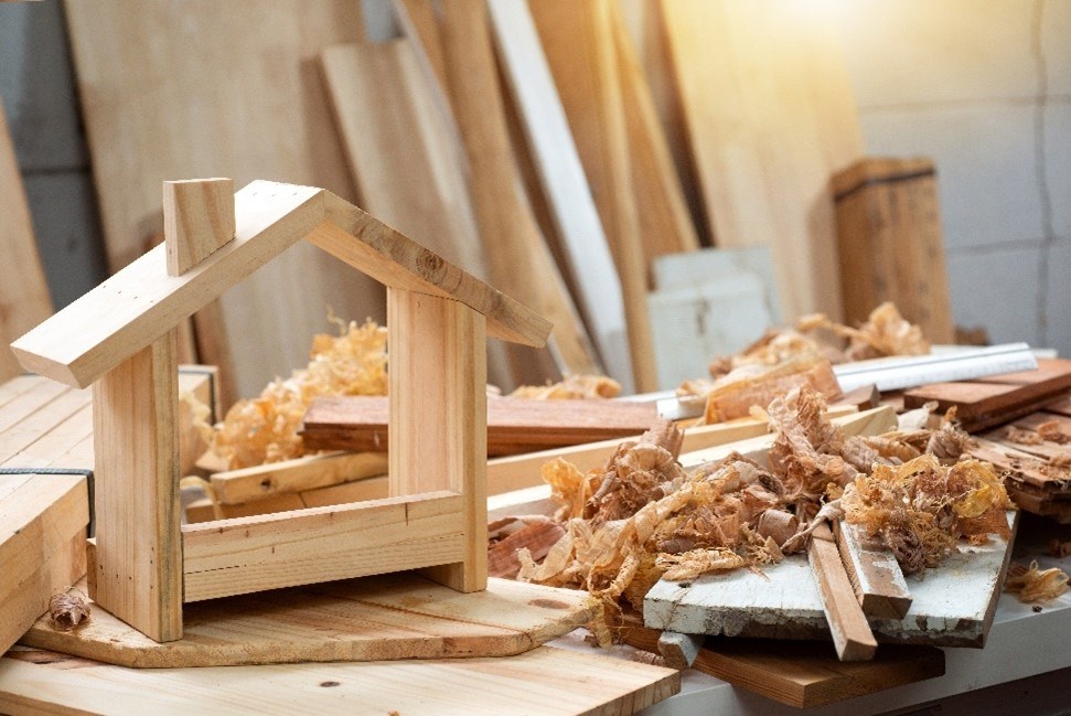 Tabletop-size house made of wood with wood scraps and sawdust next to it and planks of wood against a wall in the background