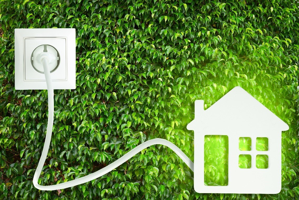 Conceptual image of an illuminated white cutout house with a cord running from it to a wall plug with a bush as the backdrop