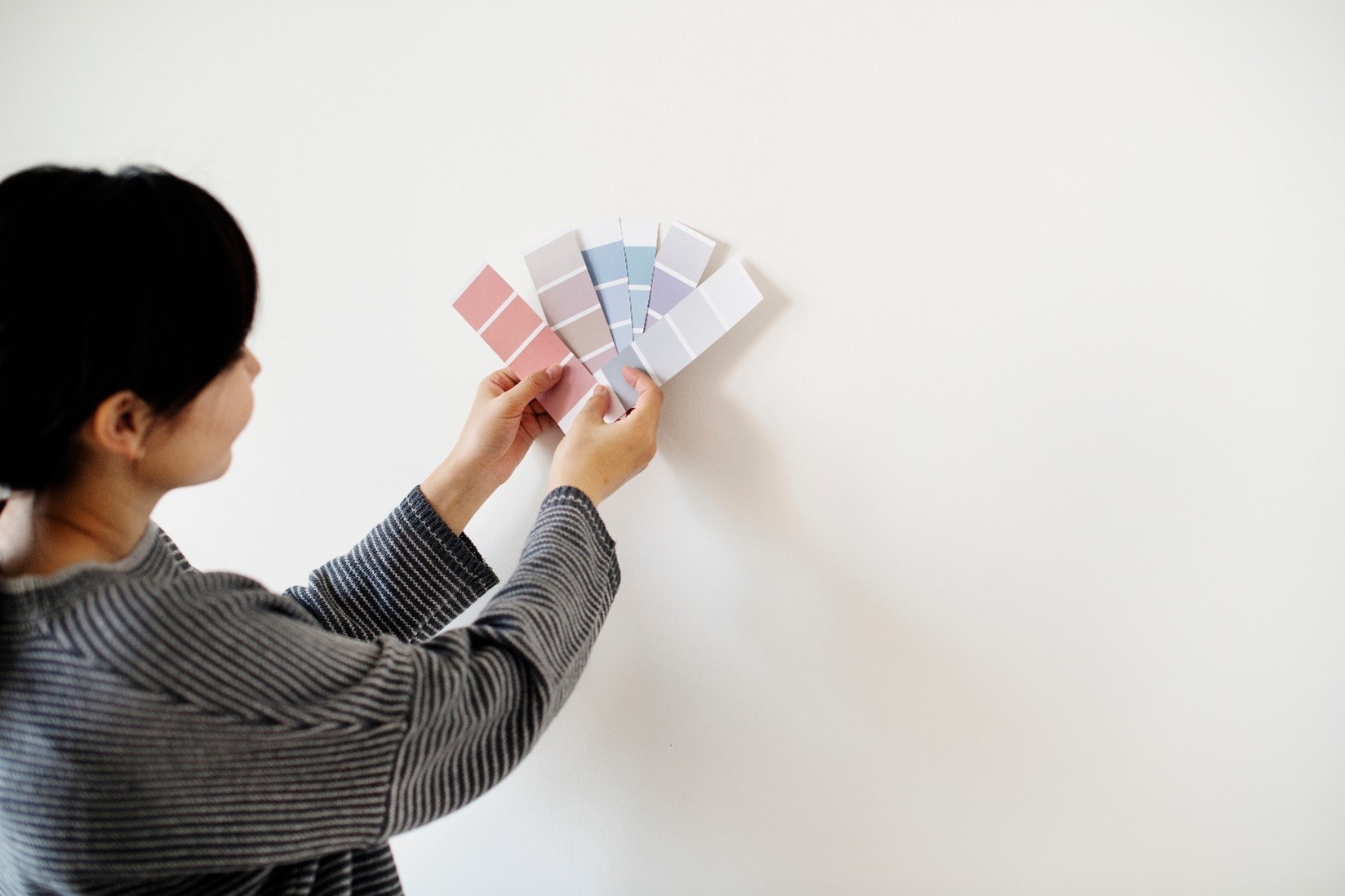 A woman in a striped shirt holds several swatches of paint colors in both hands against a white wall