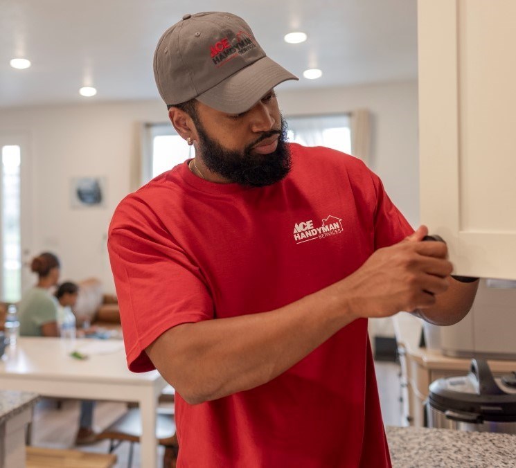 An Ace handyman wearing a red Ace shirt works on off-white kitchen cabinet doors. The home’s family sits in the background.