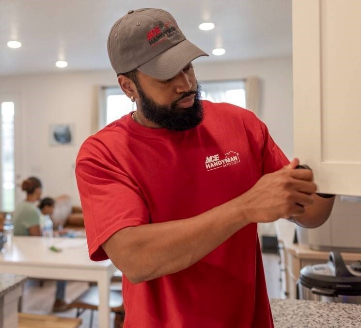 An Ace handyman wearing a red Ace shirt works on off-white kitchen cabinet doors. The home’s family sits in the background