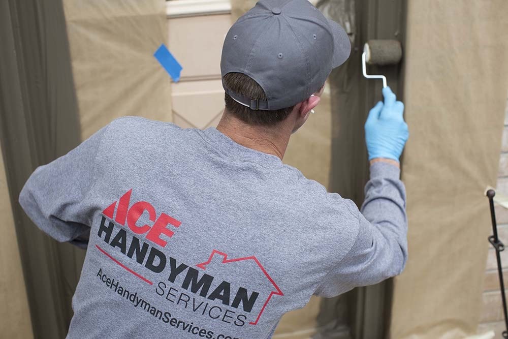 Man in a Gray Long Sleeve Shirt that Says “Ace Handyman Services Evansville” on the Back Painting a Door Jamb with a Roller