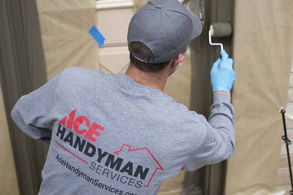 Man in a Gray Long Sleeve Shirt that Says “Ace Handyman Services” on the Back Painting a Door Jamb with a Roller