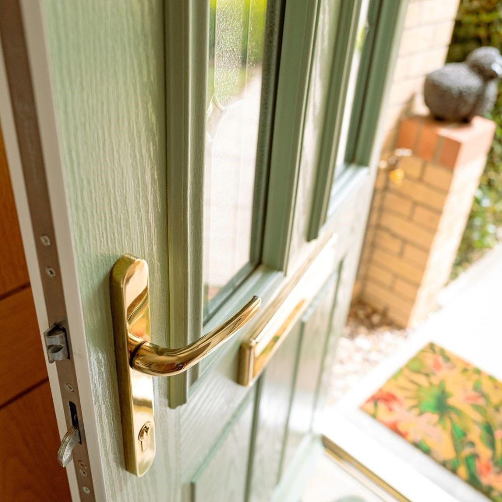 Close up of a green door opening into a home pictured from above the exterior brass lever knob with the exterior pathway and a tropical welcome mat blurry in the background. Close up of a green door opening into a home pictured from above the exterior brass lever knob with the exterior pathway and a tropical welcome mat blurry in the background.