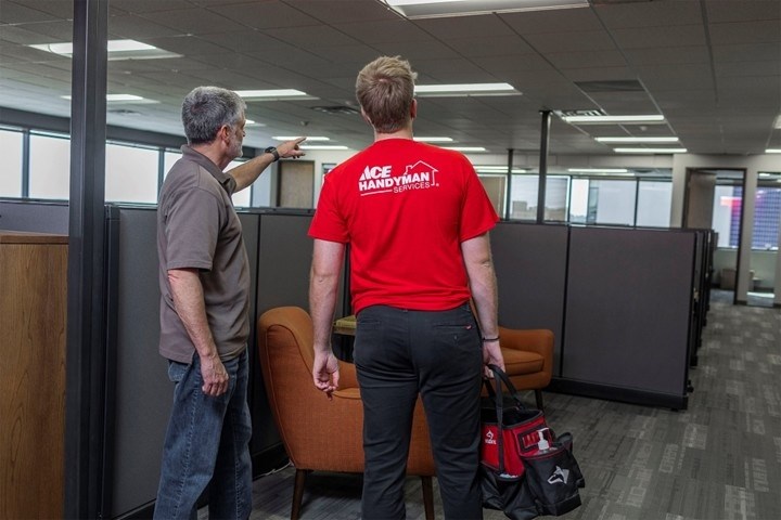 Two men standing in an office with cubicles, the one on the left points at something in the distance and talks to the other man who has on an Ace Handyman Services shirt and holds a tool bag. Two men standing in an office with cubicles, the one on the left points at something in the distance and talks to the other man who has on an Ace Handyman Services shirt and holds a tool bag.
