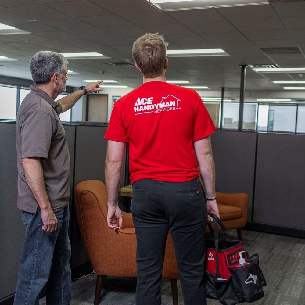 Two men standing in an office with cubicles, the one on the left points at something in the distance and talks to the other man who has on an Ace Handyman Services shirt and holds a tool bag. 
