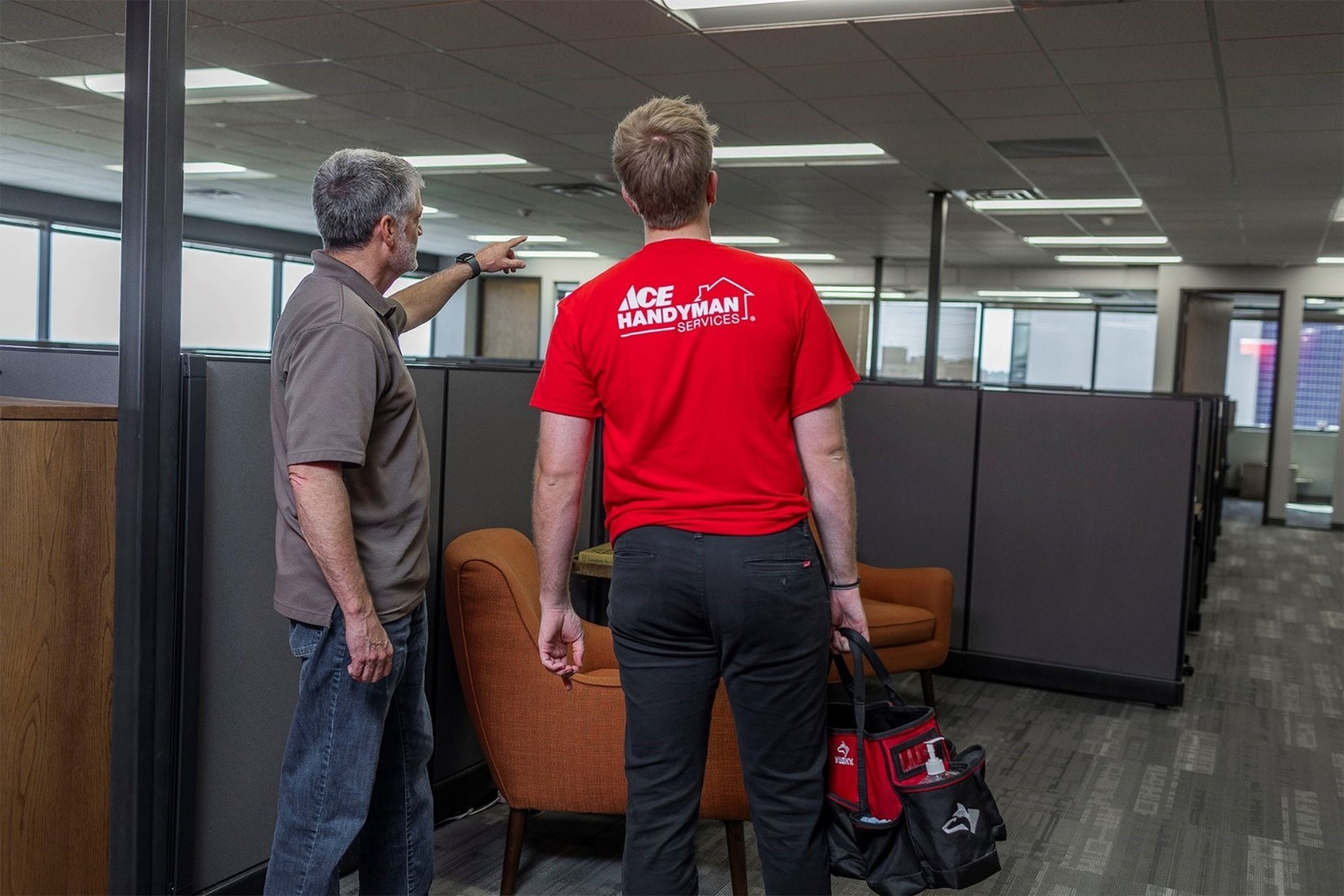 Two men standing in an office with cubicles, the one on the left points at something in the distance and talks to the other man who has on an Ace Handyman Services shirt and holds a tool bag. Two men standing in an office with cubicles, the one on the left points at something in the distance and talks to the other man who has on an Ace Handyman Services shirt and holds a tool bag.
