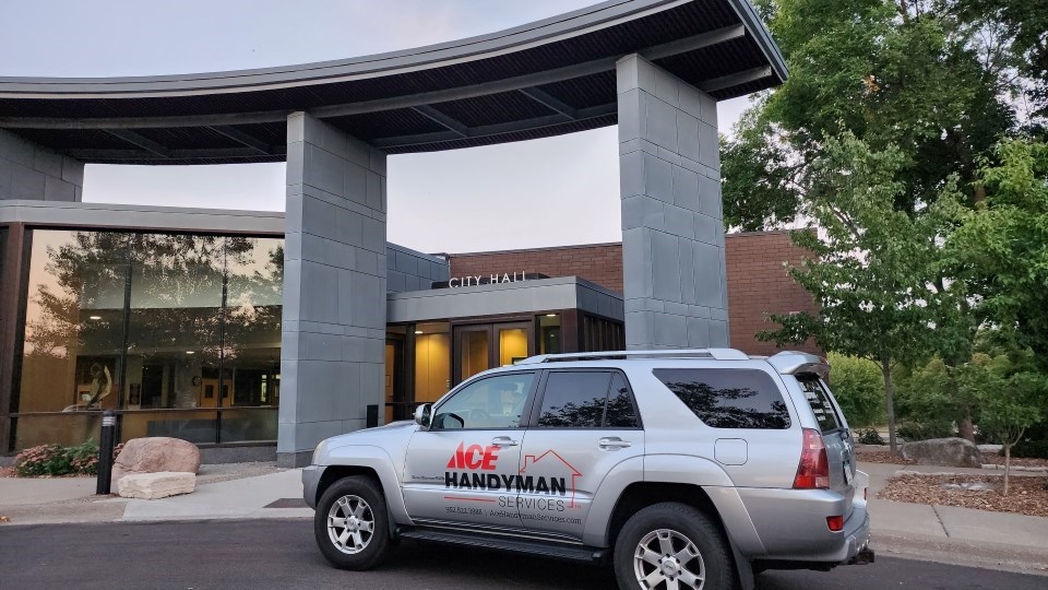 A grey Ace Handyman SUV parks outside of the local City Hall. Trees are to the right and the sun sets behind the building.
