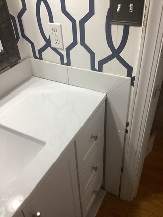 Bathroom vanity, but now the area behind the sink has been tiled with white tiles. The tile backsplash neatly covers the exposed drywall from the previous image
