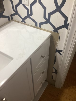 A bathroom vanity with a white countertop and a few drawers. The wall behind the vanity is painted with a blue geometric patterned wallpaper, but the area near the corner is unfinished, showing some exposed drywall.