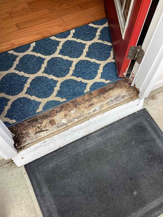A close-up of a red door with visible damage to the door frame's threshold. The wood under the door is worn and chipped, showing some exposed material.