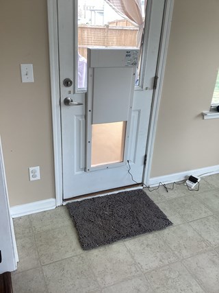 The same door, but now the doggy door frame has been installed, with a white, rectangular doggy door in place. The space in the frame is now closed with a flap, and the door is connected with a wire for an electronic mechanism.