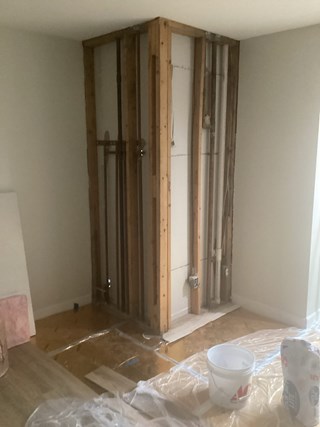 A room with a partially framed wall under construction. The structure includes exposed wooden studs and pipes, with a bucket and bag of supplies on the floor. The surrounding floor is covered with plastic sheeting, and the walls are unfinished.