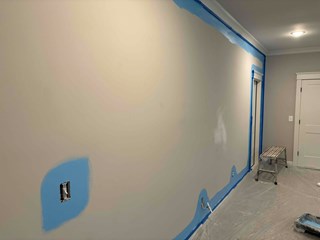 A section of a wall with the lower portion prepared for painting, with painter's tape applied to protect the edges. The wall is a neutral color, and the area around the outlet and trim is masked off. The surface is prepped, but the final coat of paint has not yet been applied.