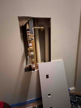 A section of drywall with a hole exposing plumbing pipes behind it.