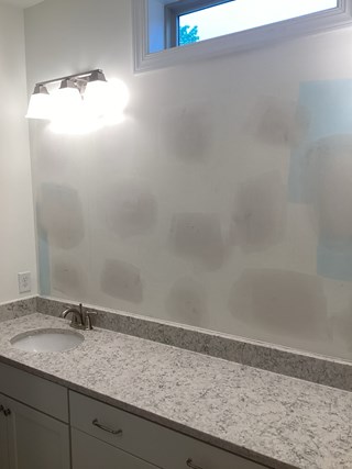 Bathroom after the repairs have been made. The broken glass has been cleaned up, and the wall now shows a freshly patched and prepared surface, likely ready for repainting.