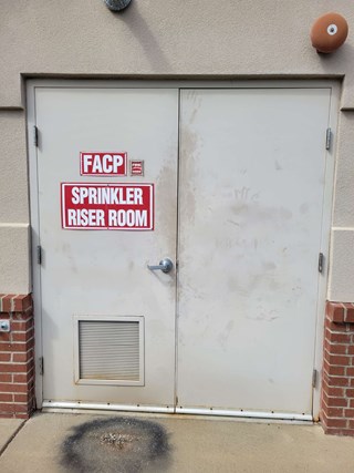 Door labeled "Sprinkler Riser Room" with visible dirt, smudges, and stains on its surface. 
