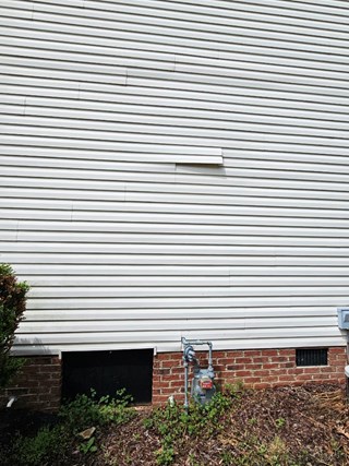 Home's exterior siding with a visible gap or damaged area, located near a vent and gas meter. The siding is misaligned, and the gap is noticeable along the wall.