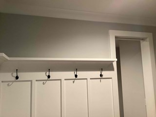 Finished area, where the drywall has been repaired. The wall now has a smooth, even surface with clean edges. Additionally, a shelf with hooks has been installed on the wall, creating a functional and polished space. The wall is freshly painted, and the area looks complete.