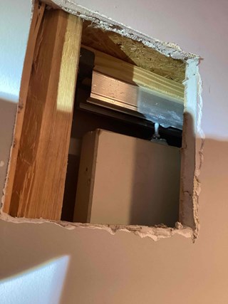 Cut-out section of drywall, revealing the wooden framing and some of the structural components behind the wall. The edges of the drywall are rough and unfinished, with visible plaster or joint compound around the opening.