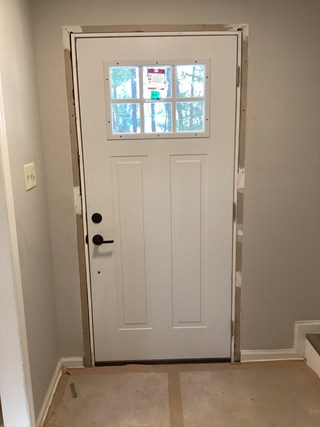 The new door is a clean white color with a window near the top. The door frame has been finished, and the surrounding wall is newly patched, giving the entire area a refreshed, neat appearance.