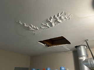Ceiling with a large hole and visible water damage. The drywall is peeling and sagging, the paint is bubbling around the hole, and the surrounding area is discolored, due to water exposure.