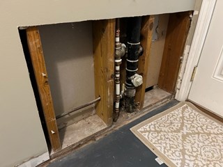 Section of wall with a large cutout revealing pipes and plumbing behind it. The drywall is removed to allow access to the plumbing