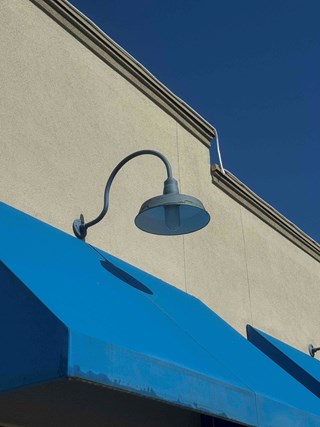 Blue commercial outdoor light fixture attached to a wall above a blue awning. The light fixture is faded, with some signs of wear and discoloration