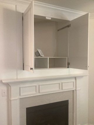 White cabinet above a fireplace with its doors open. Inside the cabinet, the space is mostly empty, except for a small shelf and an item placed inside. 