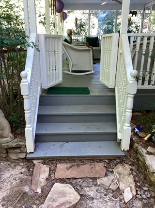 Set of stairs leading up to a porch, with wooden steps and a white railing. The steps are painted gray, but there is some visible wear and staining, particularly on the lower step.