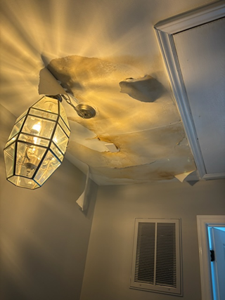 Ceiling with water damage. The drywall is sagging and peeling near the light fixture, with visible discoloration from water stains. The area around the ceiling vent also shows signs of damage.