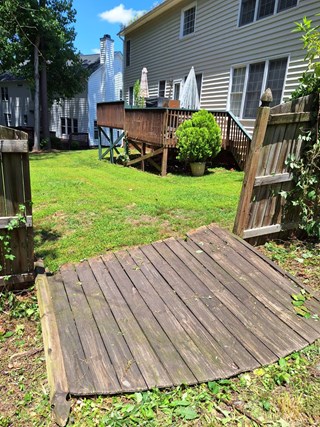 Aa wooden gate and part of a fence with significant damage, a wooden panel laying on the ground. The gate appears to be sagging and leaning to the side, and the fence planks are uneven, creating a misaligned appearance.
