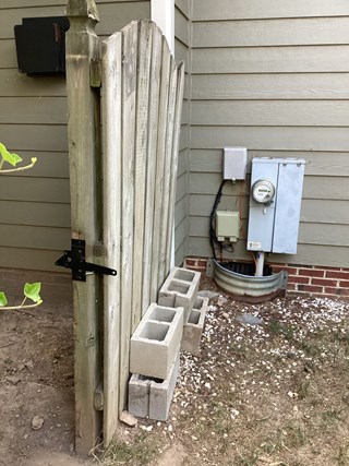 Wooden fence gate next to an electrical meter box. The gate appears to be weathered, with some leaning and damage visible. Concrete blocks are stacked next to the base of the fence, for support prior to repair.