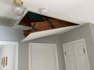 Attic access door is partially open, revealing the wooden stairs and framing inside, attic ladder arm mechanisms is broken.