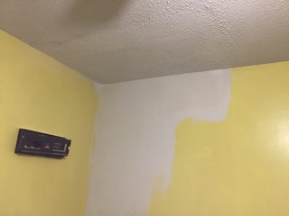 Damaged area after some work, with part of the yellow wall painted over, and the damaged ceiling area being patched and primed in preparation for further repair or repainting.