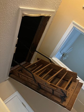 Pull-down attic ladder that is old and not fully functional, with the wooden ladder in the process of being folded down. The opening appears narrow and the ladder looks worn.