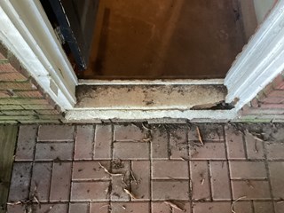 A worn and damaged threshold beneath a door, showing significant deterioration. The wood has cracked and split, and the edges are uneven. 