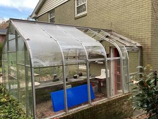 A greenhouse with clear plastic panels and a metal frame. The structure shows signs of aging and wear, with some of the panels broken and/or missing. 