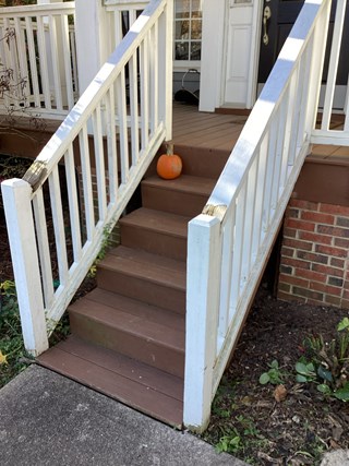 A set of worn wooden stairs leading up to a porch. The stairs have visible damage, including cracked and chipped wood. The white railing is also weathered, with signs of wear, particularly on the top edge. 