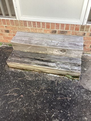 A set of weathered wooden steps leading up to a screened porch. The steps show significant wear, with discoloration, cracks, and visible damage, including one of the steps being uneven and falling apart. 
