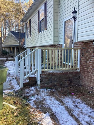 A front porch renovation with a new wood railing and white steps. The porch is brick-paved and leads to a walkway,