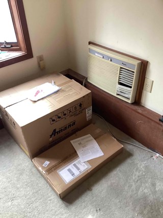 Old air conditioner being replaced – A new Amana in wall AC unit box is seen on the floor of a room, with the existing air conditioner unit soon to be removed.
