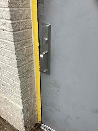 nstallation of a commercial-grade door handle on a restaurant exterior door in Cary, NC. Shows the handle, lock cylinder, and attachment plate, with brickwork and a yellow safety strip visible.
