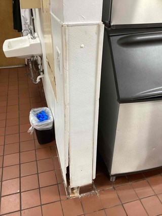 Before photo of a damaged wall corner in a commercial kitchen in Raleigh NC 27610. The wall covering is torn and broken, revealing the wall structure. A sink, ice machine, trash can, and tiled floor are also visible.