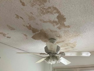 : Before photo of a ceiling with a large water stain in Raleigh, NC 27609. The stain is irregularly shaped and brownish, indicating potential water damage. A ceiling fan is visible below.
