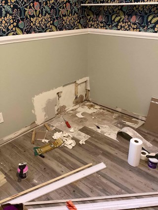 Before photo of drywall damage in a room in Garner, NC. Hole in the wall with exposed studs, wallpaper above, and various spots of flooring damaged. 