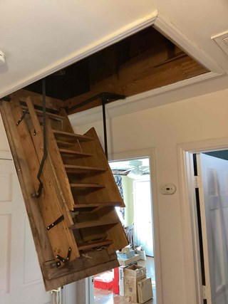 Before photo of an older, wooden attic ladder in Raleigh NC 27609. The ladder is partially extended, showing signs of wear and age. A cluttered attic space is visible through the access hatch.