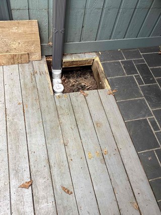 Close-up of a damaged deck with a missing section of decking boards, creating a hole. Shows weathered gray wood and a gray downspout. Location: Cary, NC 27511. Prior to repair by Ace Handyman Services.