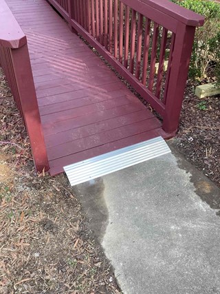  After photo of a ramp transition in Raleigh, North Carolina, showing a red stained wooden ramp with a newly installed silver metal threshold plate leading to a gray concrete surface, improving accessibility.