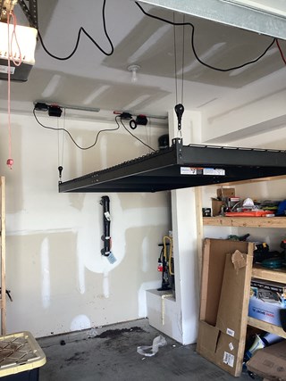  A garage with a completed overhead storage rack installation. The black metal rack hangs from the ceiling, supported by cables and pulleys